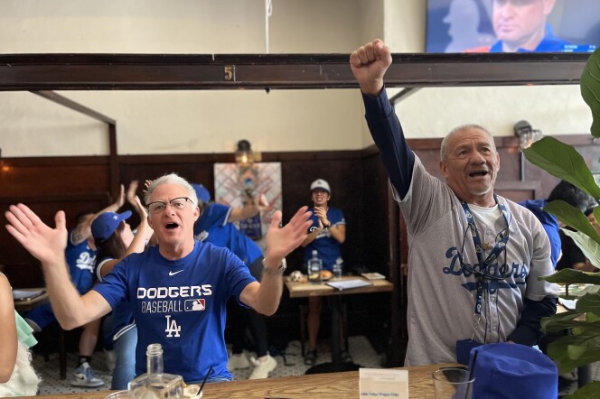 A white male wearing a blue Dodgers shirt and a Latino man wearing a gray Dodgers shirt raise their arms in excitement.