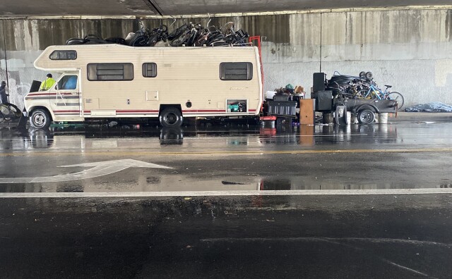 An RV is park under a freeway overpass with bicycles piled on top of it and on a trailer behind it.
