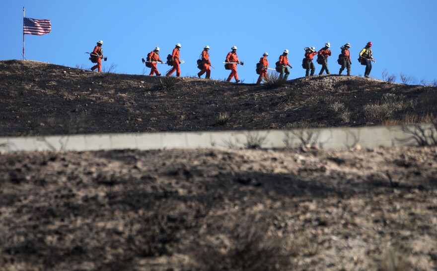 Firefighters in orange jumpsuits walk along a ridge with a U.S. flag waving