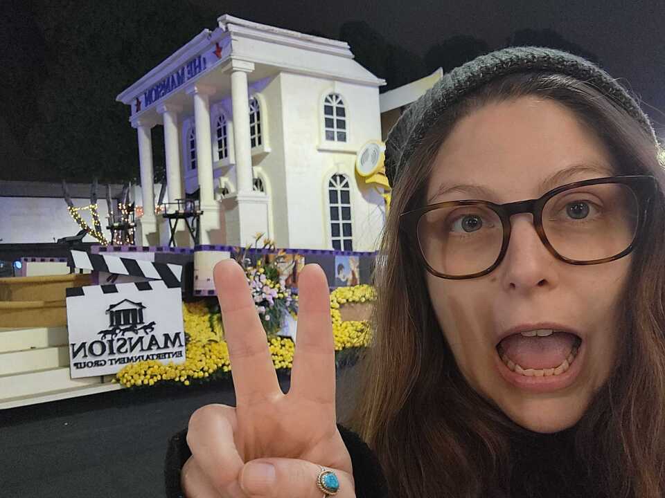 A woman with glasses holds up a victory sign in front of a parade float