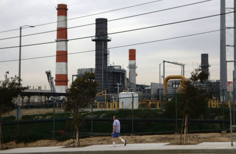 Red and white smoke stacks rise behind power lines and a person walking on a sidewalk