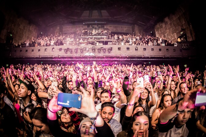 A theater auditorium is filled with people for a performance. They're all looking towards the camera... some have their hands up as if they're dancing, others are smiing and whooing