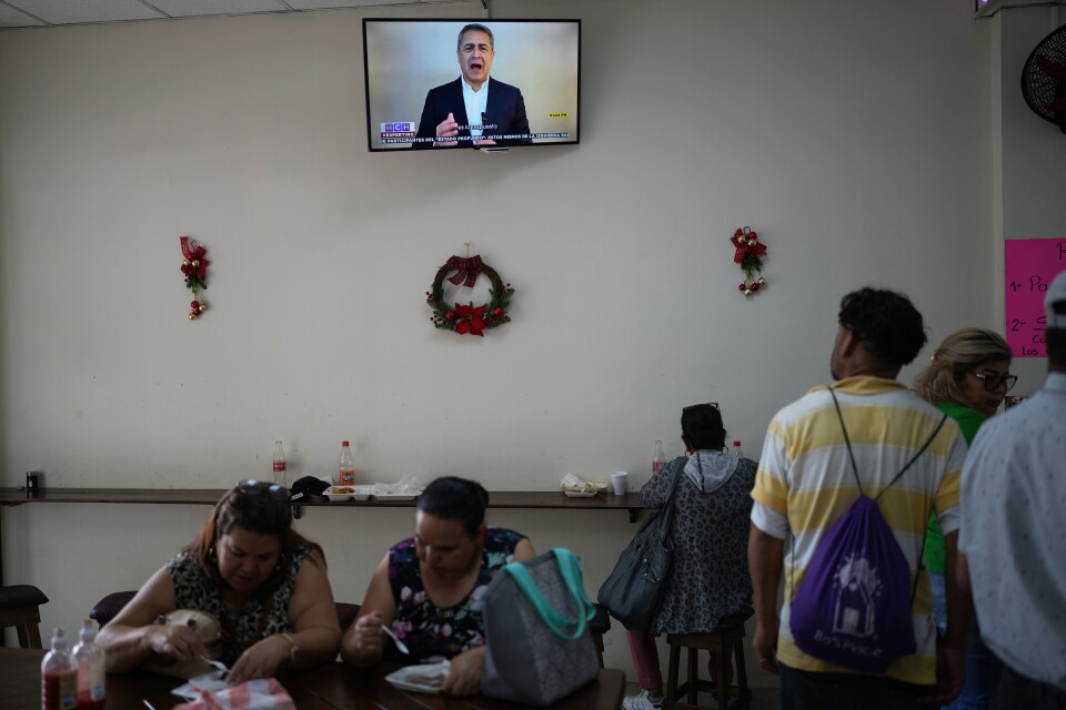 A man speaks on a TV screen on a wall that has sparse wreath decorations.