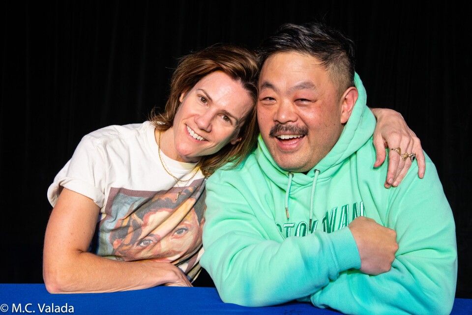 Cameron Esposito, a white woman wearing a t-shirt and Chris Grace, a dark-skinned man with a mustache wearing a green hoodie, smiling at the camera