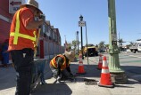There are two men in high-visibility vests and wide-brimmed hats on a road. One is standing next to a blue wheelbarrow with a shovel. The other man is on his knees using a tool to spread wet concrete over box where street lighting wires are stored. 