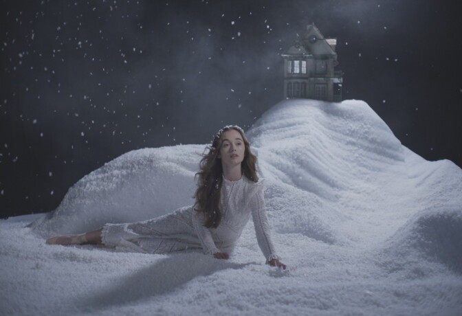 A woman in a white gown lies on a white snow-covered stage scene with a house in the background. 