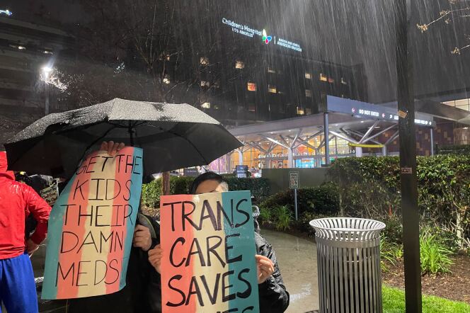 Two people hold up umbrellas and protest signs with pink, white and light blue signs to signify the transgender pride flag. The signs say "Trans care saves lives" and "Get the kids their damn meds."