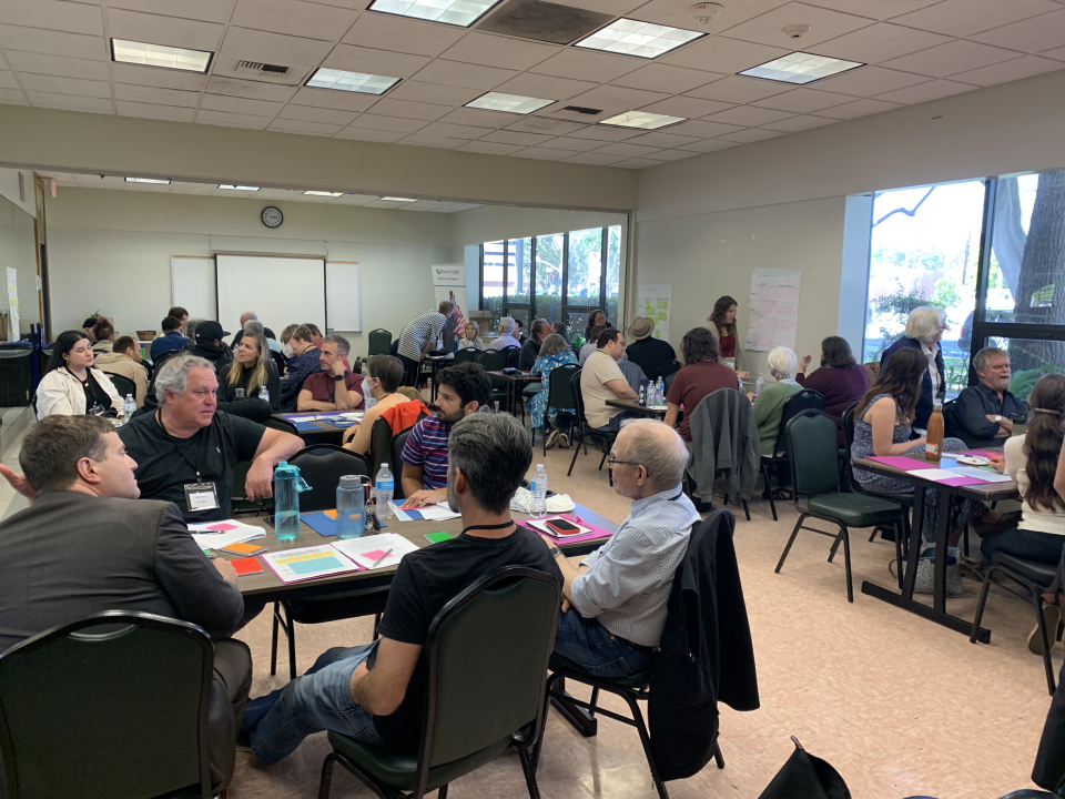 Several groups of people sit at tables full of papers and colored sticky notes in a recreation center.