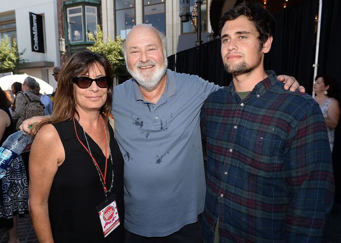 Director Rob Reiner, a man with light skin tone, bald head and white beard, smiles as he stands in between and hugs his wife, Michele Singer, a woman with light skin tone, wearing a black dress and sunglasses, and son, Nick Reiner, a man with light skin tone, short goatee, wearing a dark-colored flannel. They pose for a photograph with Rob Reiner and Michele Singer look at the camera, while Nick Reiner looks away.