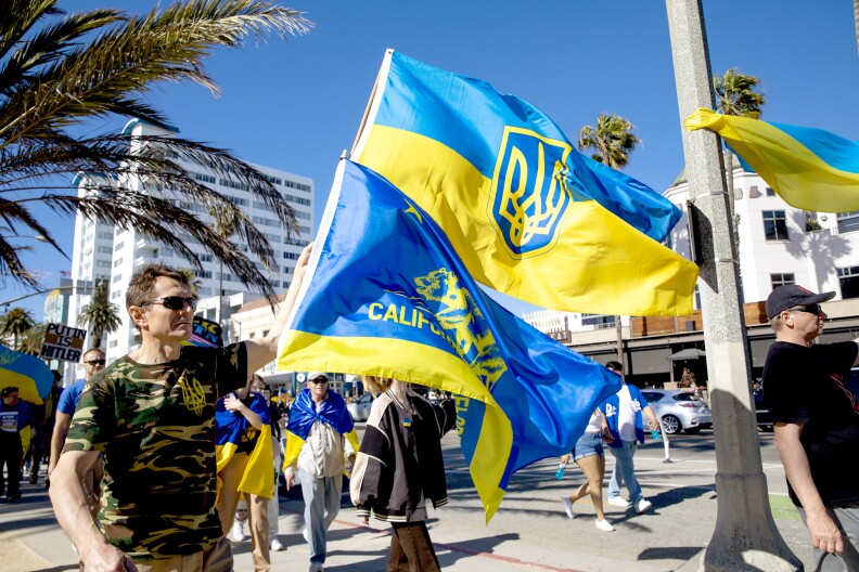 A crowd of about eight people walks on the sidewalk along a road in Santa Monica. A caucasian man at the front of the photo on the left holds two flags, one a version of the state of California's flag, but colored Blue on the top eighty percent and gold on the bottom twenty percent in the style of the Ukrainian glag. The bear and California republic logo on the flag is also gold. The second flag is the Ukrainian flag, half blue on top, half gold on bottom, with a blue and yellow shield logo in the center. The man wears a green, tan, and brown camouflage t-shirt with a gold cursive script logo on the top right corner. Behind the man are two people wearing blue and gold Ukrainian flags draped over their shoulders. White buildings sit in the background on the other side of the street. A man deep blurred in dark shadow in the background holds a sign which reads "Putin Is Hitler" in black text on a white background.