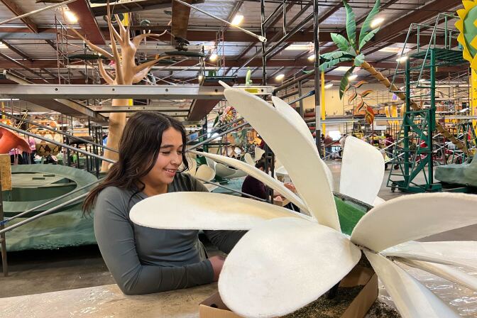 A girl with dark brown and medium-light skin tone holds a petal of a giant white flower.