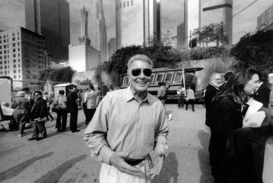 A black and white wide view of a man with a light skin tone and dark pilot shades on smiling in at the camera. He's standing neara crowd of people and a small train car next to the downtown L.A. skyline.