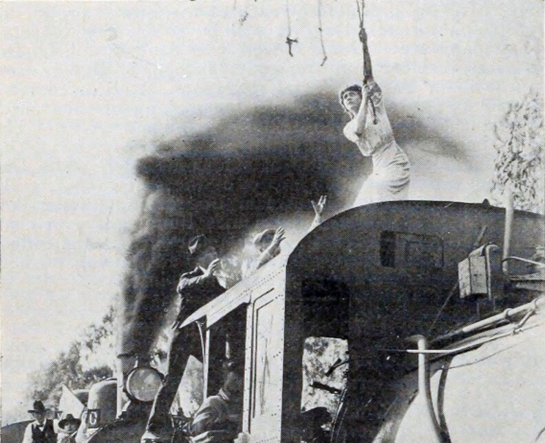 A black and white photo of a woman standing on top of a train as black smoke billows upwards, holding onto a rope above her head. Two men stand below with their arms out.