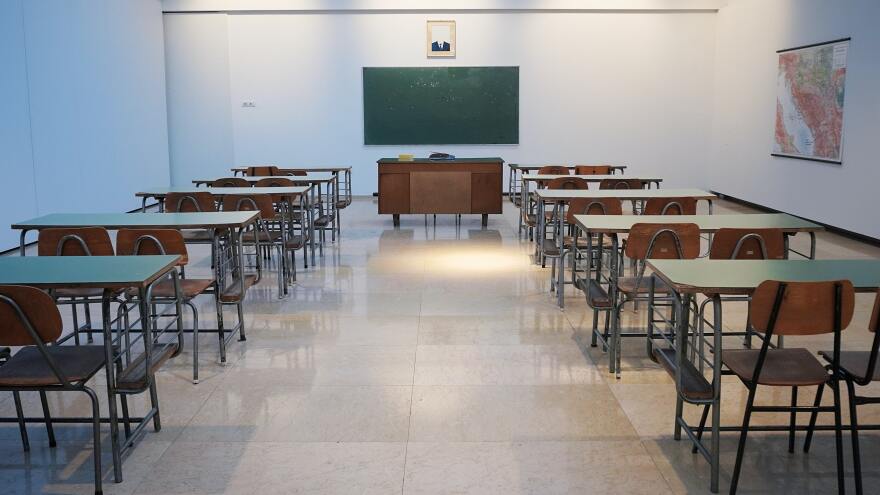 An image of an empty classroom with two rows of desks. At the front of the room there's a chalkboard and a desk in the middle.