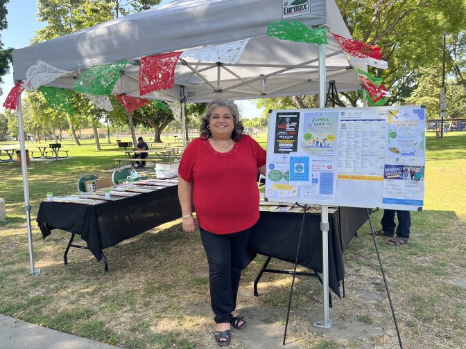 A woman with medium skin and short hair is standing in front of her information booth at a park in southeast Los Angeles.
