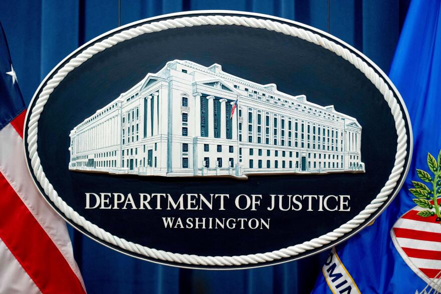 The seal of the Department of Justice - an oval shaped seal with an illustration of a large white building in the middle. It hangs on a wall between two flags