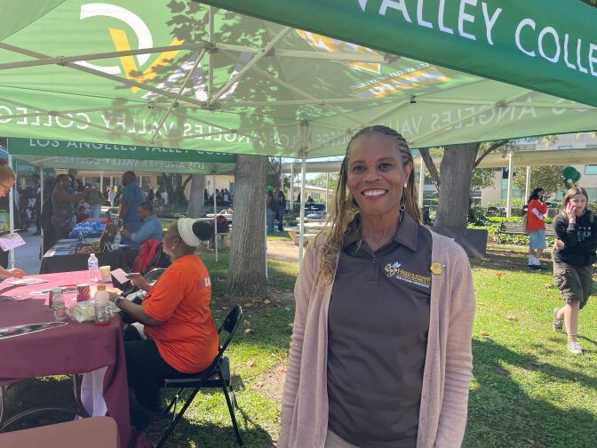 A woman wearing a brown collared shirt and cardigan stands in the foreground under a canopy tent. 