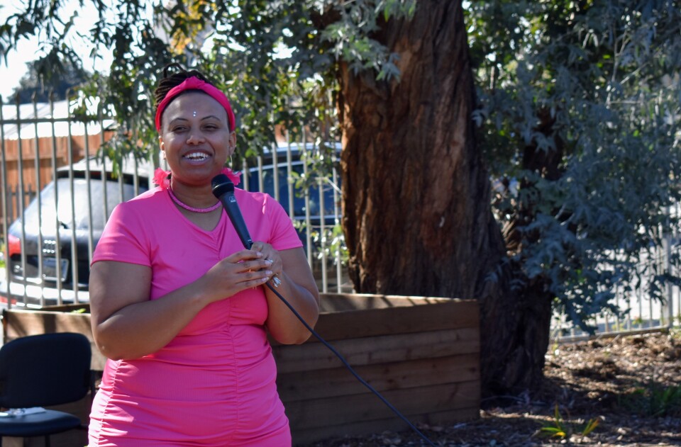 A woman with dark-toned skin woman wearing a dark pink T shirt and pants, with a pink headband and necklace, talks and smiles into a microphone. She stands next to a tree.