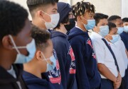 Adolescent boys, most of whom are Black or Latino, stand in a line with face masks, school sweatshirts or white button-downs and ties. They are shown from the side. 