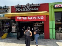 The storefront of La Azteca Tortilleria, a well-known Mexican eatery. A large red and white banner reading "WE'RE MOVING" is prominently displayed across the yellow awning above the entrance, signaling that the business is relocating. Two women stand out front,  waiting to enter. 