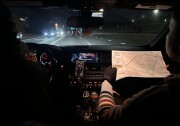 The interior of a car from the vantage point of the backseat. The person on the right in the passenger seat is holding up a printed map with both hands, which are wearing fingerless black gloves. The interior is slightly illuminated by buttons on the dashboard, with streetlights and car headlights shining through the front windshield. 