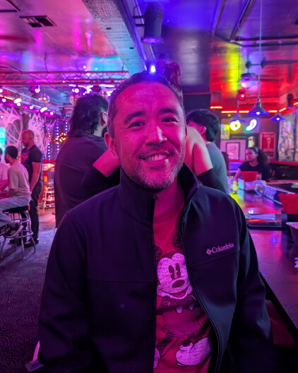 An Asian man with a buzz cut and a Mickey Mouse shirt sitting at the bar.