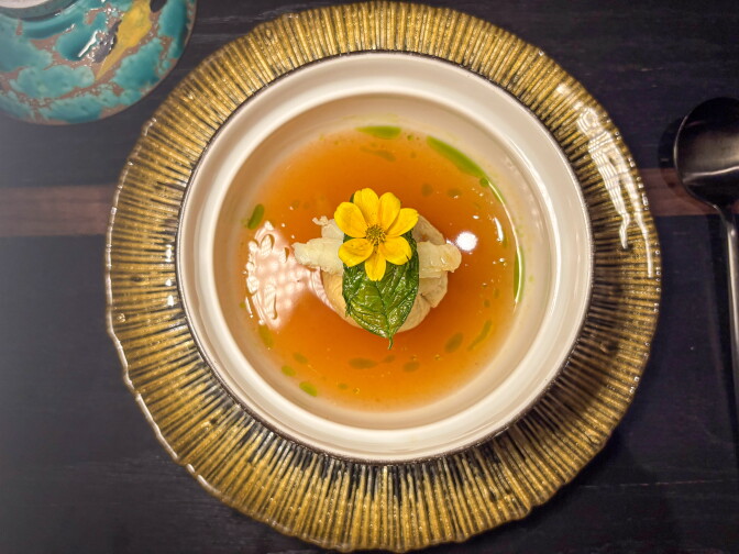 Overhead view of a white bowl of bright orange broth on a gold-rimmed plate. A small bundle of crab and shrimp in the center is topped with a green leaf and a yellow flower. Green oil is dotted across the broth.