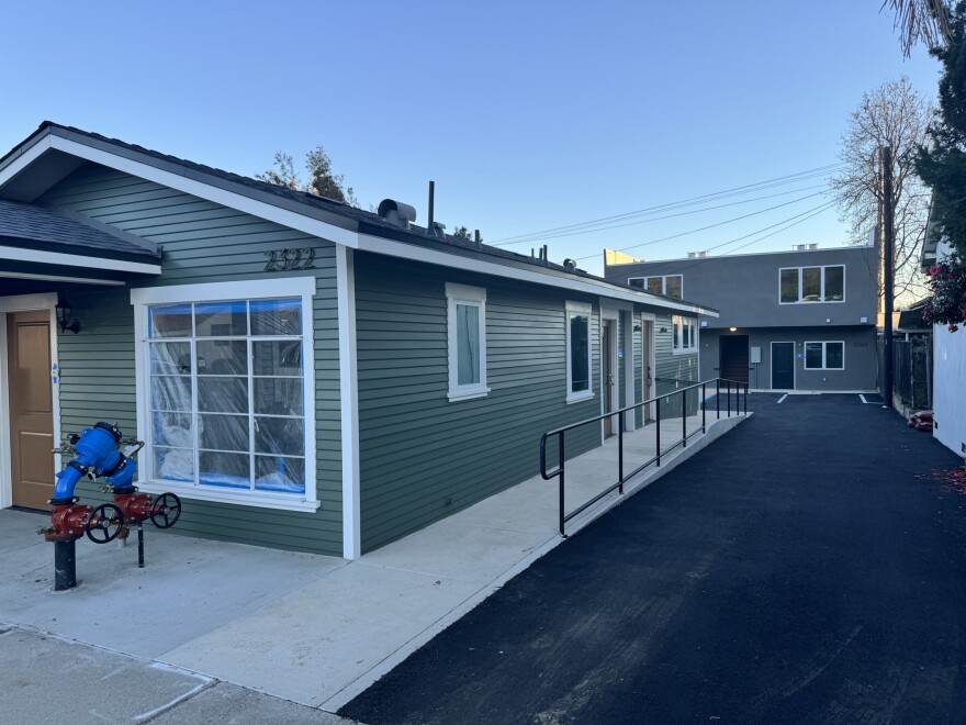 A one-story green building with newly-installed windows sits in the foreground and another two-story complex sits behind. 