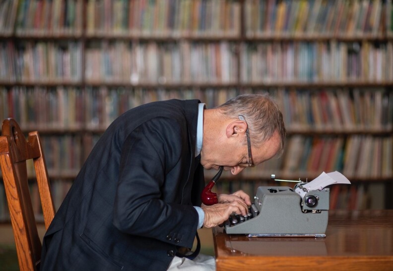 A side shot of David Sedaris with a pipe in his mouth, leaning over an old manual typewriter. Books on shelves are in the background.