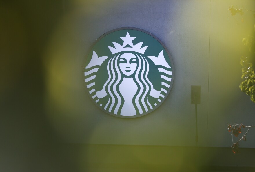 A close up of the Starbucks logo, which is an illustration of a siren in green and white. There are plants in the foreground of the photo.