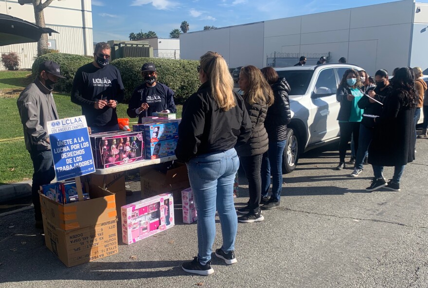 People stand outside of Jon Donaire's building for a toy drive. There are toys on a table, and a sign that reads "Unete a la lucha por los derechos de los trabajadores."