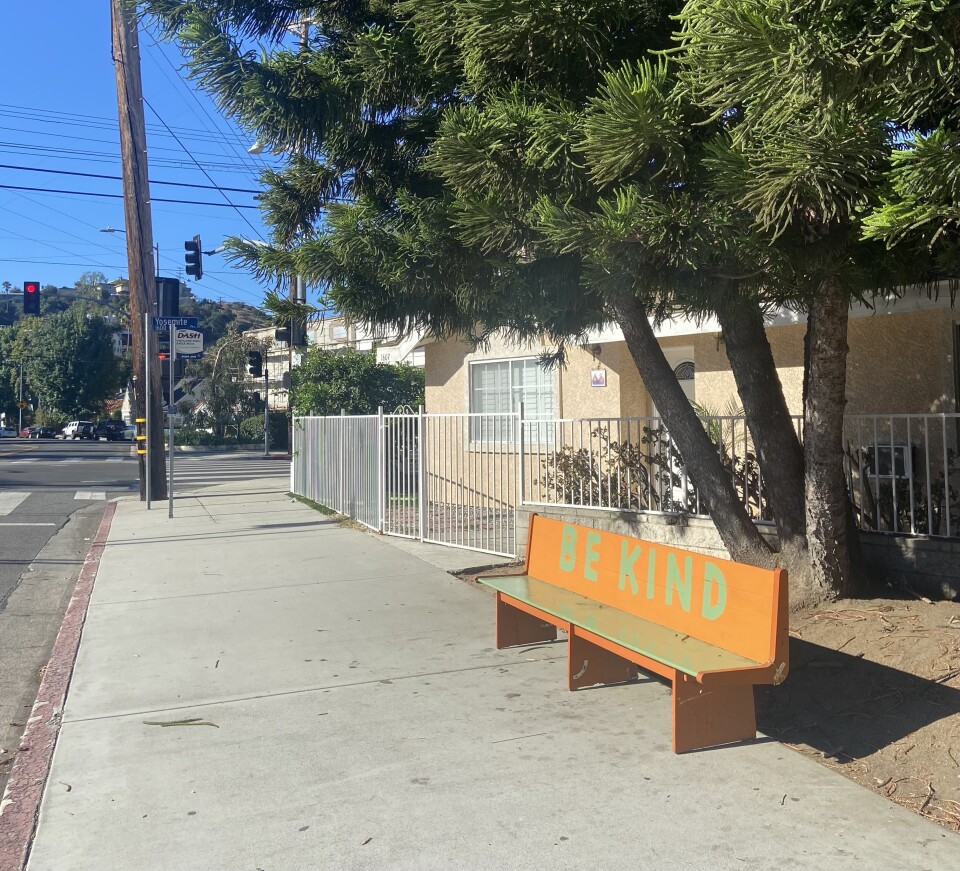 A bench, possibly a church pew, with the words "Be Kind" in large green font on a sunny sidewalk near a bus stop.