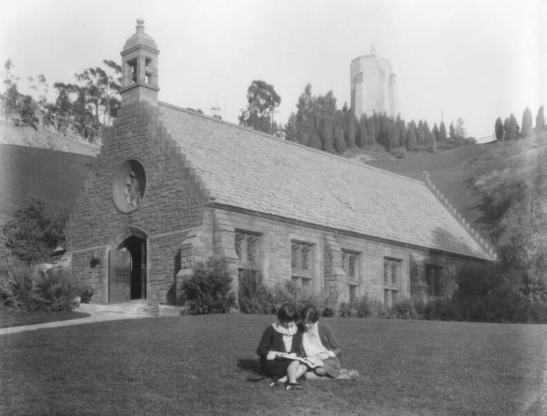 A black and white photo of two women sitting on a lawn in front of a large, one-story stone chapel. They are looking at what appears to be a book. In the background on a hill in the distance, is a large white tower.