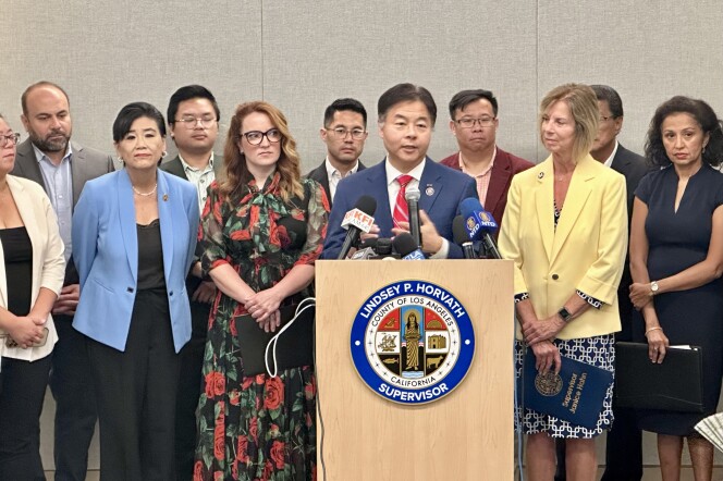 An Asian American man in his 50s speaks at a podium, flanked by ten men and women on both sides of him. 