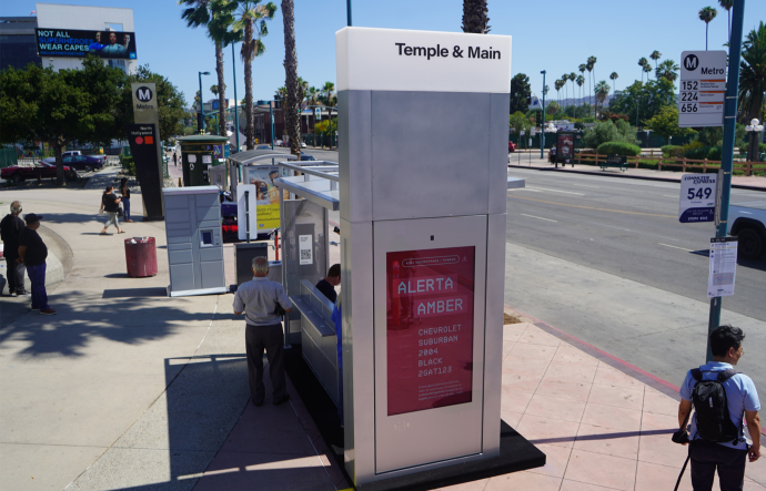 A person stands in the shade provided by a bus shelter as other people stand and walk around at a transit station next to a street.