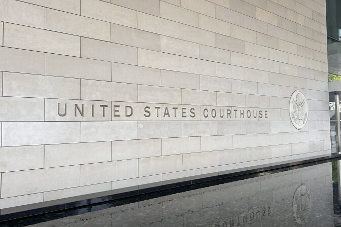The exterior beige stone exterior has the words "United States Courthouse" engraved with the American federal seal beside it. The letters are reflected from a stone water fountain below the stone exterior wall at the federal courthouse in downtown Los Angeles. 
