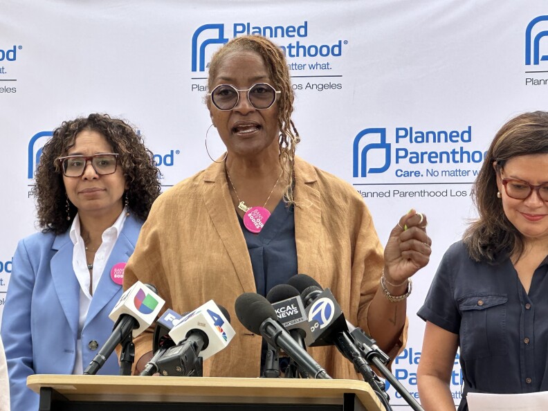 A woman speaks passionately at a podium during a Planned Parenthood Los Angeles event, flanked by two women. She wears large round glasses, a light brown jacket, and a pink “Bans Off Our Bodies” button. Multiple microphones from news outlets, including Univision, NBC 4, and ABC 7, are positioned in front of her. The backdrop features the Planned Parenthood logo and slogan.