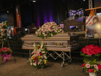 Large flower arrangements sit on and near Valentina Orellana Peralta's coffin. A large photographs of her stands on easels on either side of the coffin. A drum set is on a stage in the distant background.