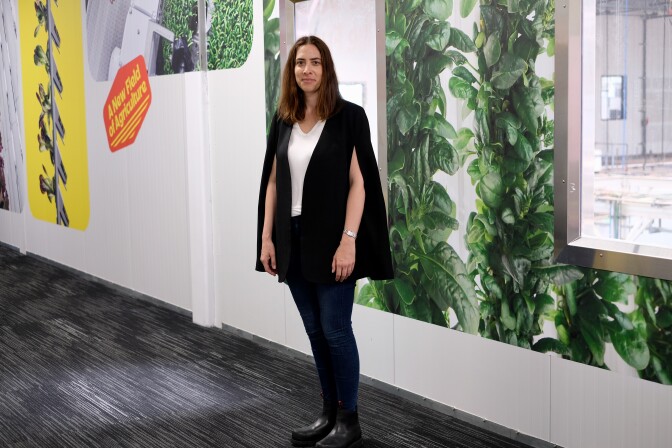 A white woman with shoulder length straight brown hair wears a black shawl over a white shirt and jeans and black boots. She's centered in the frame and the background is a white wall with windows and large images of leafy greens. 