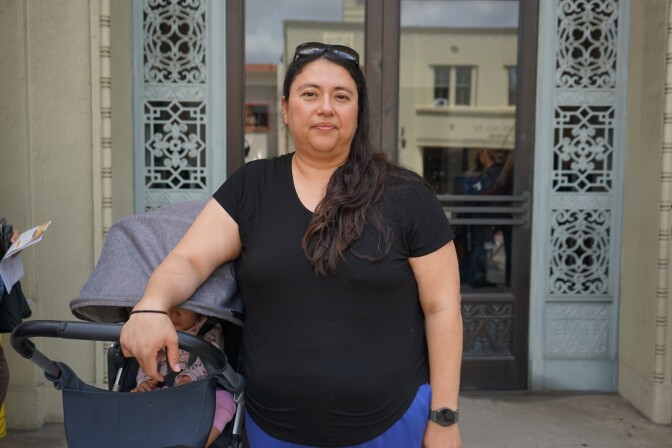 A woman with a medium skin tone wearing a black shirt and blue pants stands in front of a glass door with ornate metal lining on either side. She rests her right arm on a gray stroller with a baby inside.