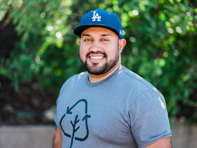 A headshot of a man with light brown skin wearing a blue LA dodgers hat and grey shirt with a recycling motif on the front. THe man smiles and has a black short beard and mustache. The background is blurred greenery.  