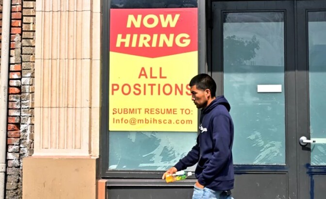 A man carrying a water bottle and wearing a blue hooded sweatshirt walks past a storefront with a sign hanging up that says, "Now hiring, all position" with information on where to send a resume.