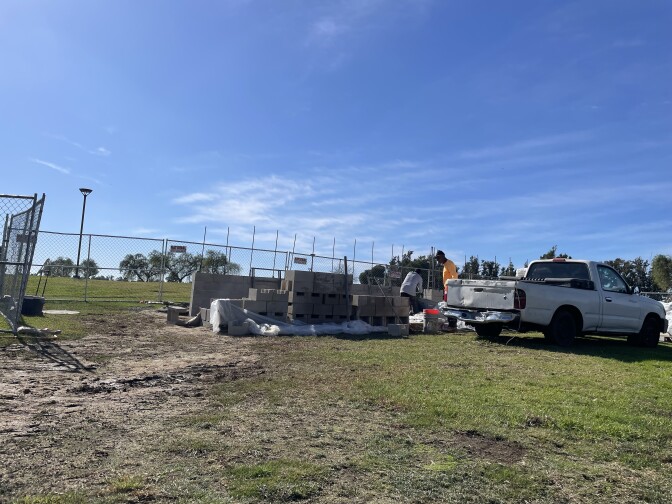A wide show showing two men among a pile of cement blocks next to a white pickup truck. There's fencing in the back. It's a blue-sky day in a grassy and muddy field. 