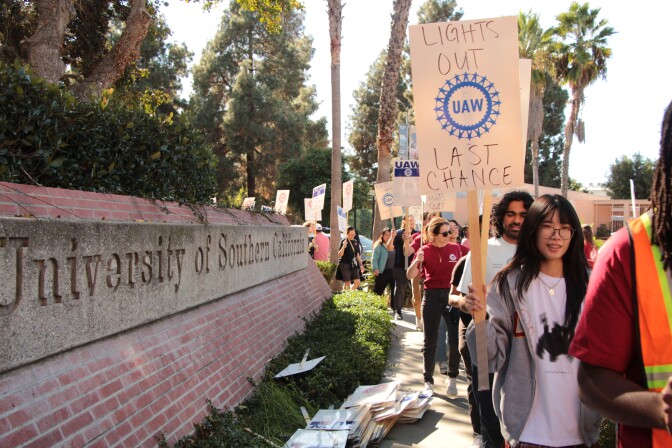 People hold picket signs and walk by a wall with the words "University of Southern California"