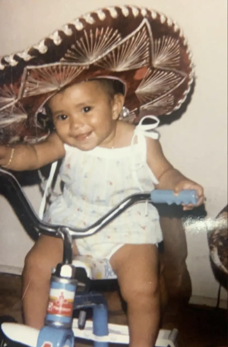 Flor at age three sits on a tricyle in a one-piece outfit that ties at the shoulders. She's wearing a big sombrero.