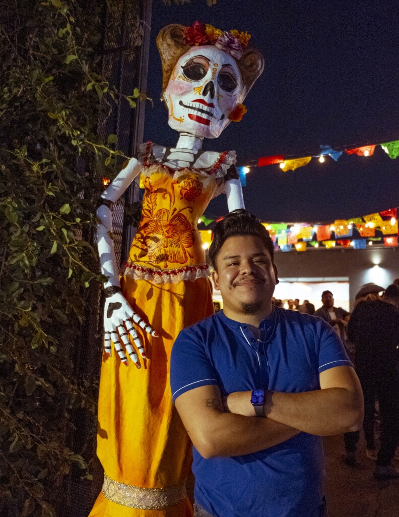 A portrait of Kevin, a man with a medium skin tone, as he stands in front of a large catrina that's wearing an orange dress. He's smiling at the camera while wearing a blue t-shirt. Bright papel picado hangs in the background.
