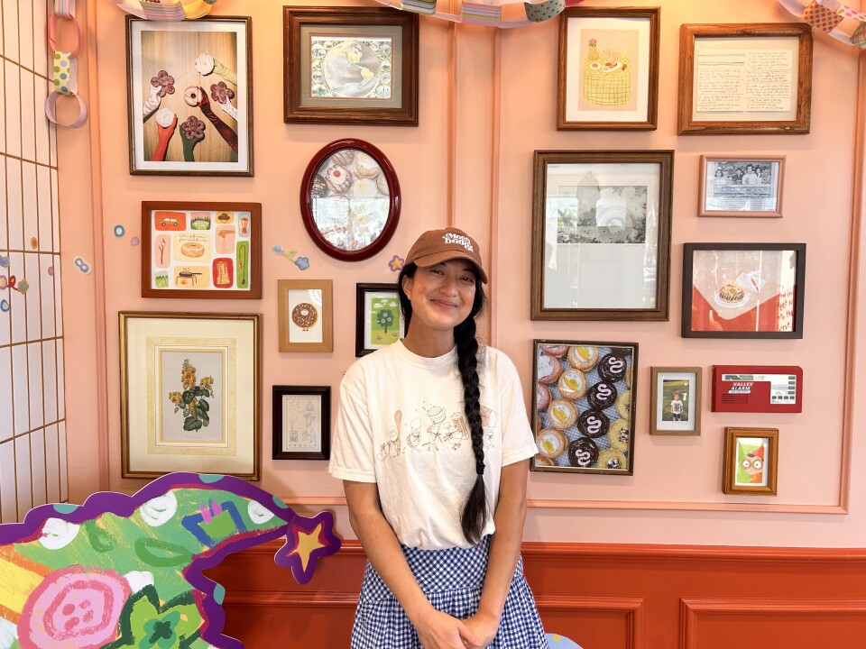 An Asian woman with her long black hair resting in a braid over her shoulder is smiling at the camera in front of a light pink wall full of framed pictures. Her hands are gently clasped in front of her. She's wearing a brown hat, a white t-shirt, and a blue and white checkered skirt. 