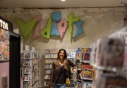 A feminine presenting person with light skin tone and light brown hair wearing a brown leather jacket and jeans stands leaning on a stand full of magazines. Behind them on the wall is a colorful sign that reads "Vidiots."