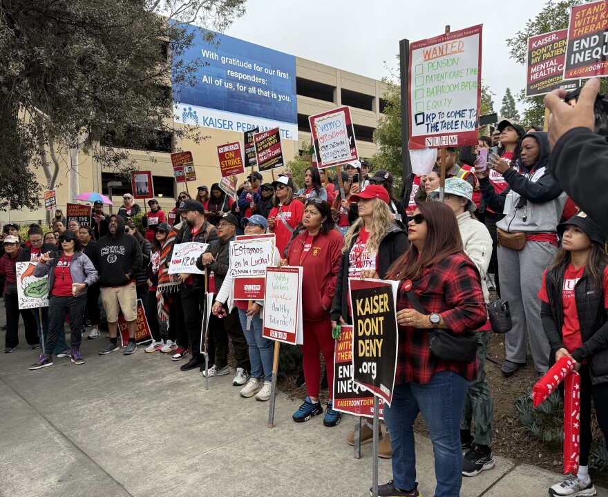 A group of dozens of striking mental health workers and their supporters stand at a rally in Pasadena. They mostly wear red colors and hold protest signs against Kaiser.
