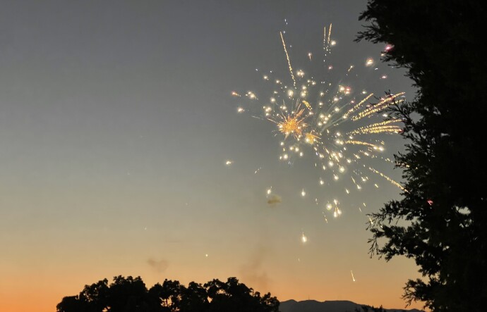 A firework sparkles silver, yellow and orange at sunset, with trees and hills framing the image.